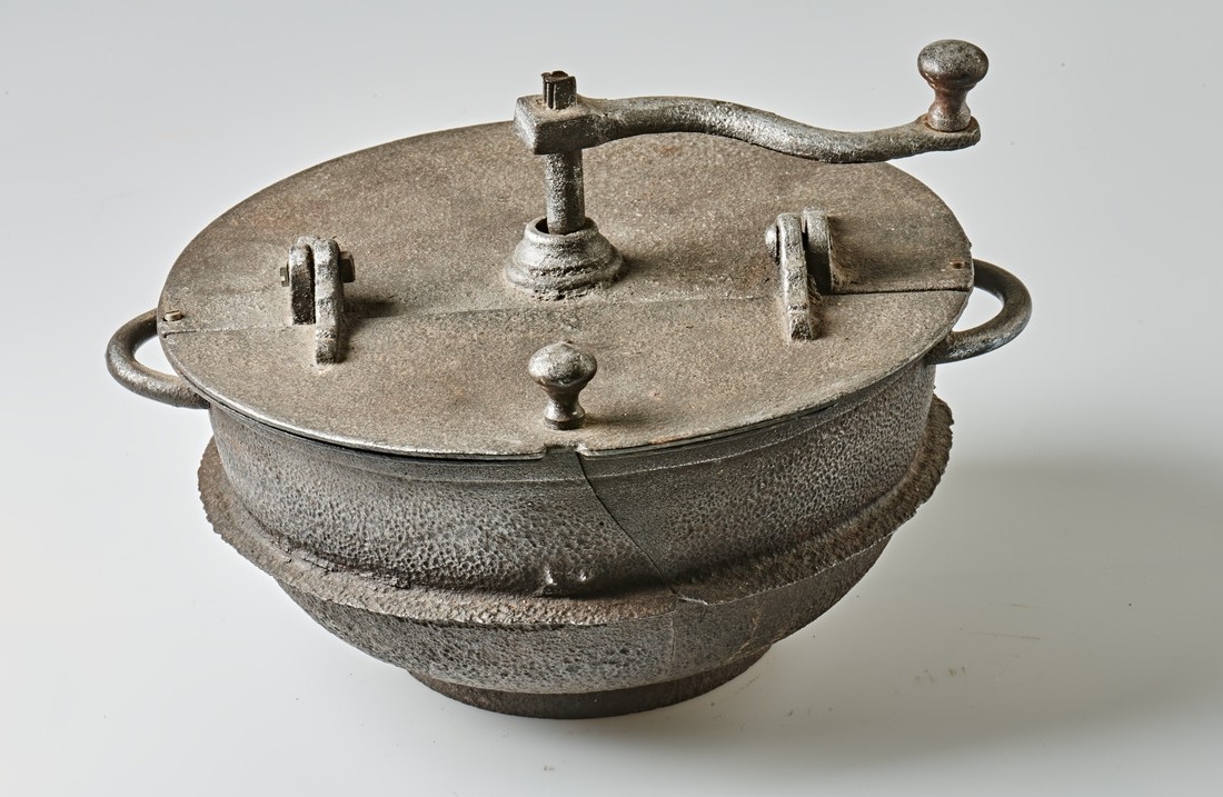 Coffee roaster without a handle, made of iron, to be inserted in an economy stove. The lid is divided in the middle, one half can be opened via two hinges for filling or removing the roasted coffee beans. There is a small knob on the outside for better handling. A handle is attached to the other half of the lid for regularly turning the beans during roasting. The body of the roaster has a small handle on both sides.