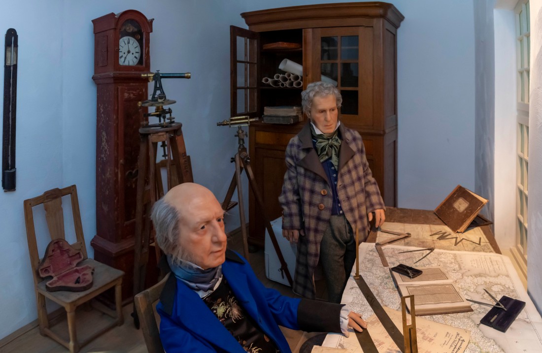 Zwei Männer vor einem Schreibtisch mit Zeichenblättern, Lineal und anderen Messwerkzeugen.