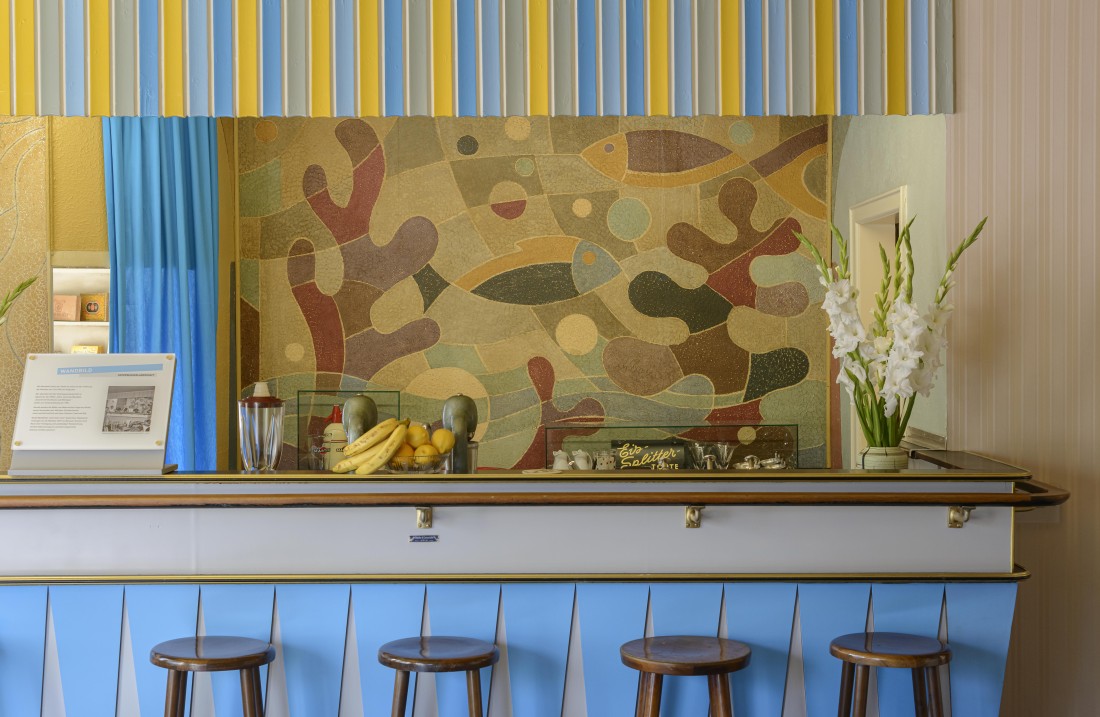 light blue clad counter, in front of it 4 wooden stools. Behind the counter murals with green plant-like motifs