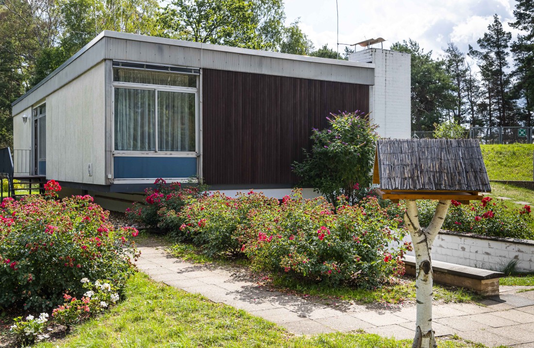 Quadratisch wirkendes Haus mit weißer Seitenwand und Front mit Fenstern und brauner Verkleidung, im Vordergrund ein Vogelhäuschen