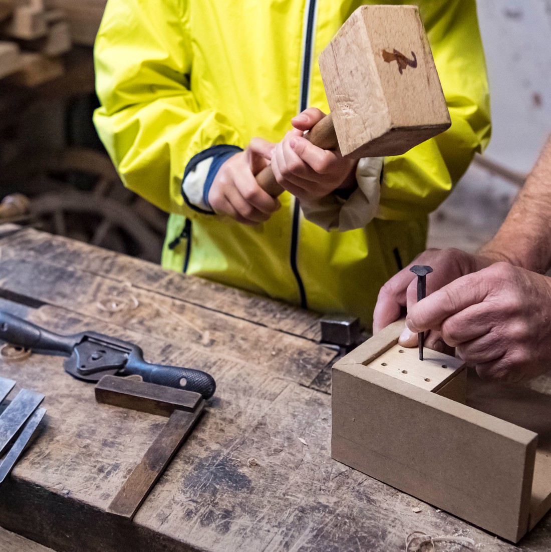 Kind schlägt mit Holzhammer auf einen Nagel, der von einem Mann auf einem Holzstück gehalten wird