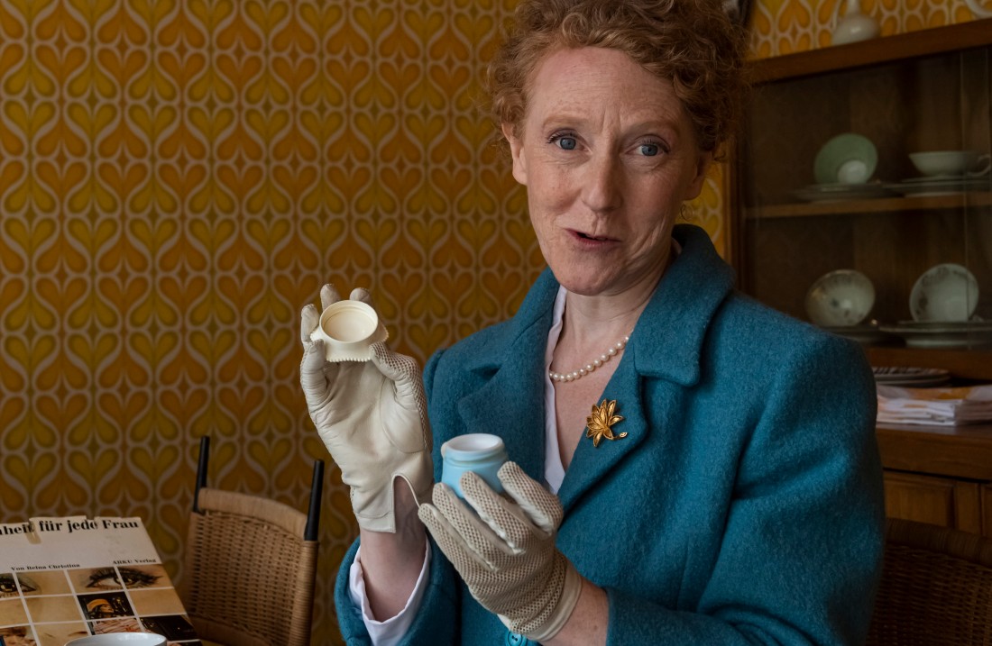 A woman in a blue suit sits at a table with many small boxes and leafs through a notebook