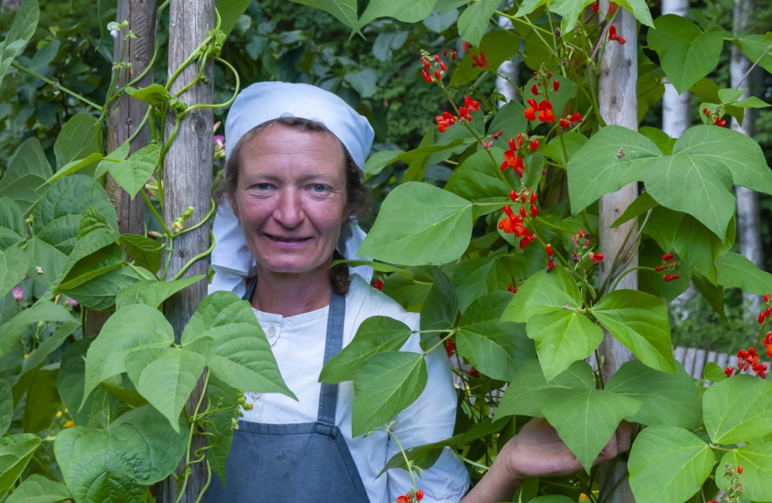 Eine Frau mit weißem Kopftuch steht zwischen Büschen mit großen Blättern und roten Blüten
