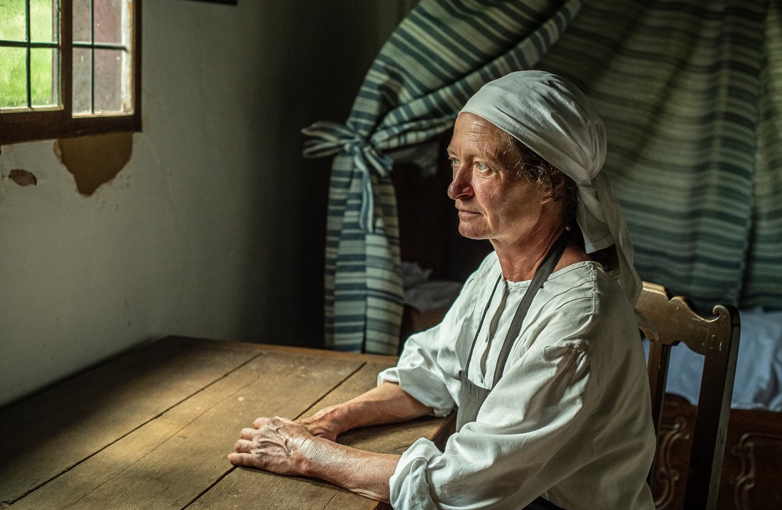 Frau mit weißem Kopftuch sitz an einem Holztisch und schaut aus dem Fenster.