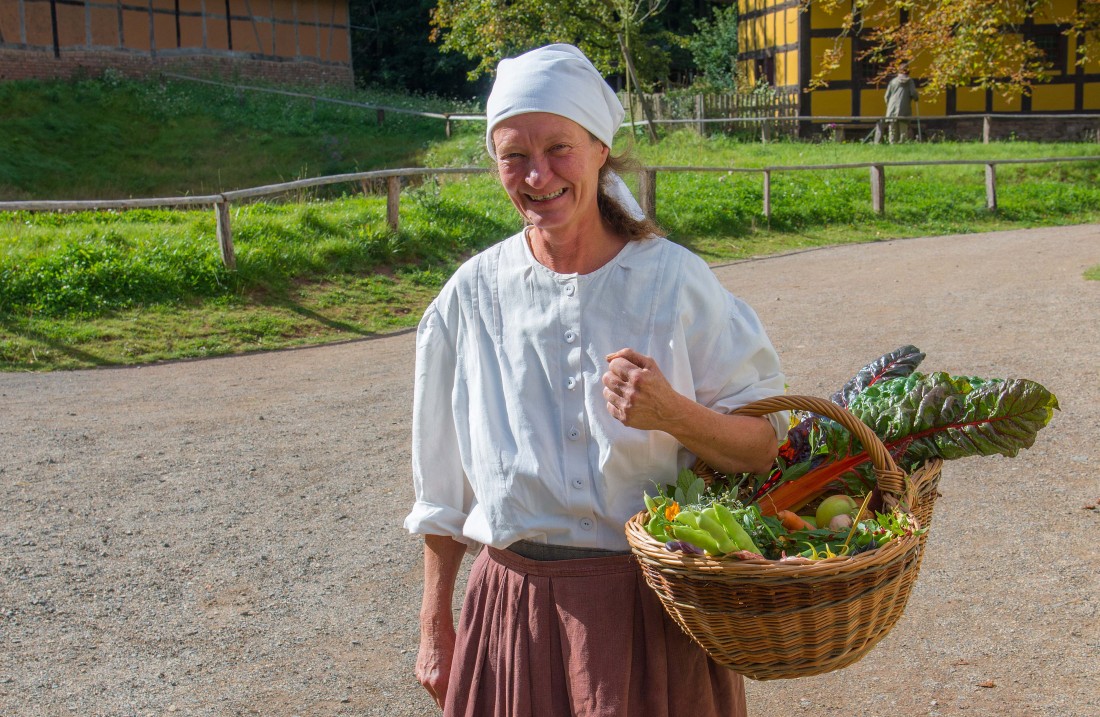 Frau mit weißer Bluse und Kopftuch geht mit einem Korb voller Gemüse über dem Arm einen Weg entlang.