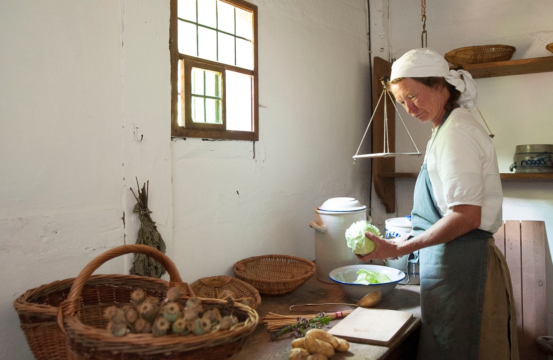 Woman stands in front of a table with potatoes and cabbage and holds a cabbage in her hand to cut it