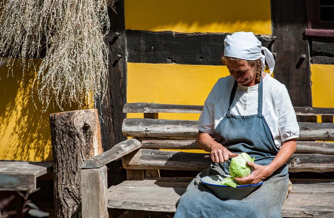 A woman with a white headscarf sits on a bench in front of a half-timbered house and cuts a cabbage.