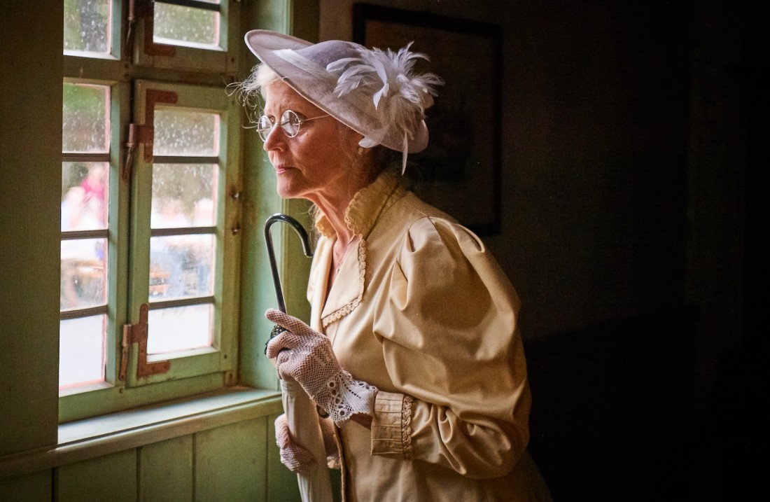 Lady in a hat and blouse is holding a parasol and looking out the window