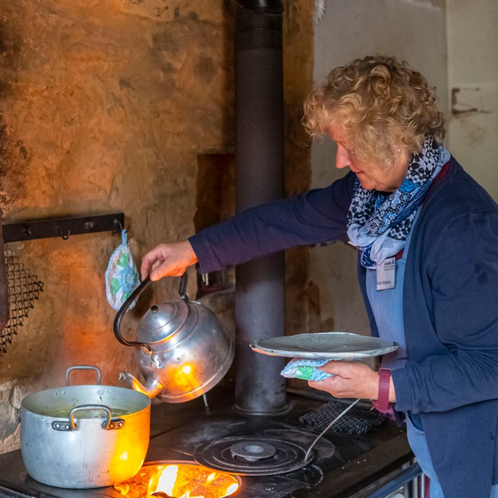 Eine Frau gießt aus einem Teekessel heißtes Wasser in einen Topf. Dieser steht auf einem Sparherd, unter der Herdplatte sieht man das Feuer brennen.