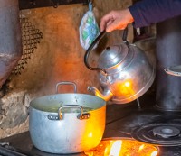 Eine Frau gießt aus einem Teekessel heißtes Wasser in einen Topf. Dieser steht auf einem Sparherd, unter der Herdplatte sieht man das Feuer brennen.