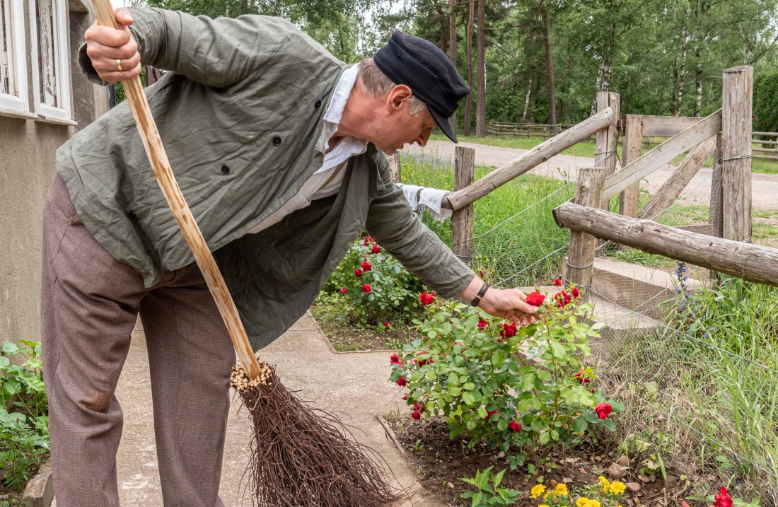 Mann hält einen Besen in der Hand und bückt sich zu einer Blume im Beet.