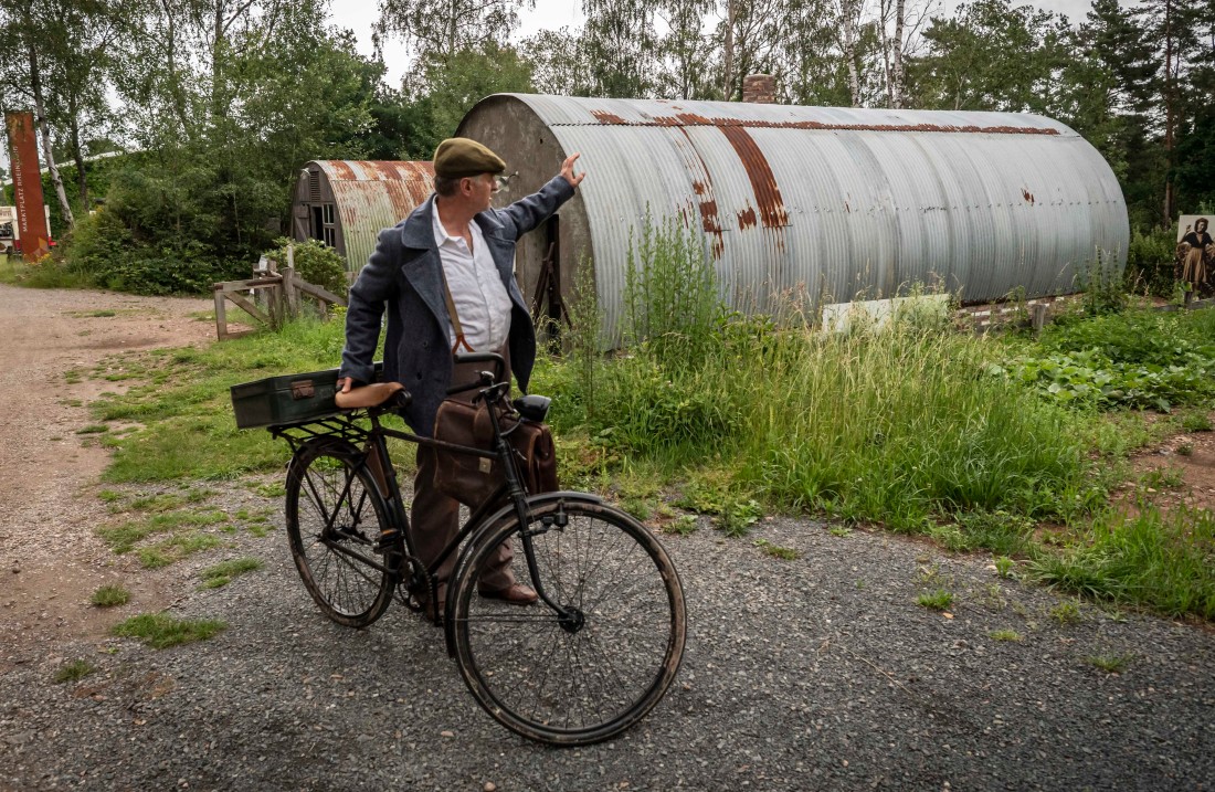 Mann steht neben seinem Fahrrad und winkt Frau hinter einer Wellblechhütte zu