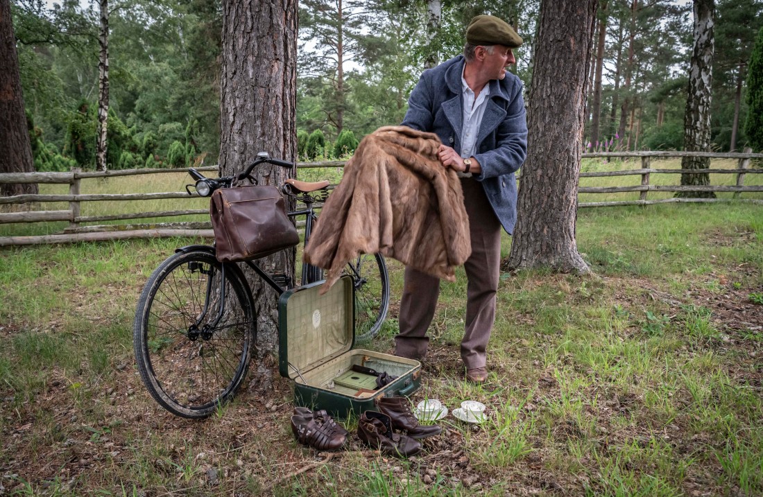 Mann hält einen Pelzmantel in der Hand, ein Fahrrad mit Tasche am Lenker steht neben einem Baum und ein Koffer mit kleineren Objekten liegt vor ihm im Gras.