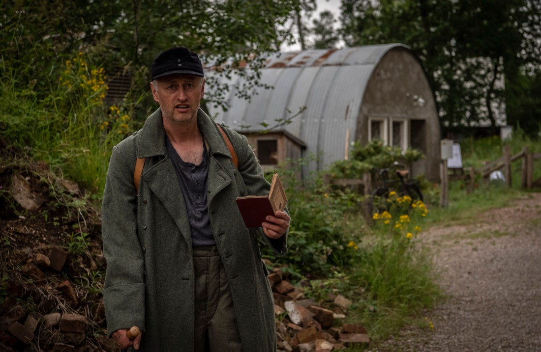 Mann mit Armeemantel hält einen Pass in der Hand und steht vor einer Nissenhütte