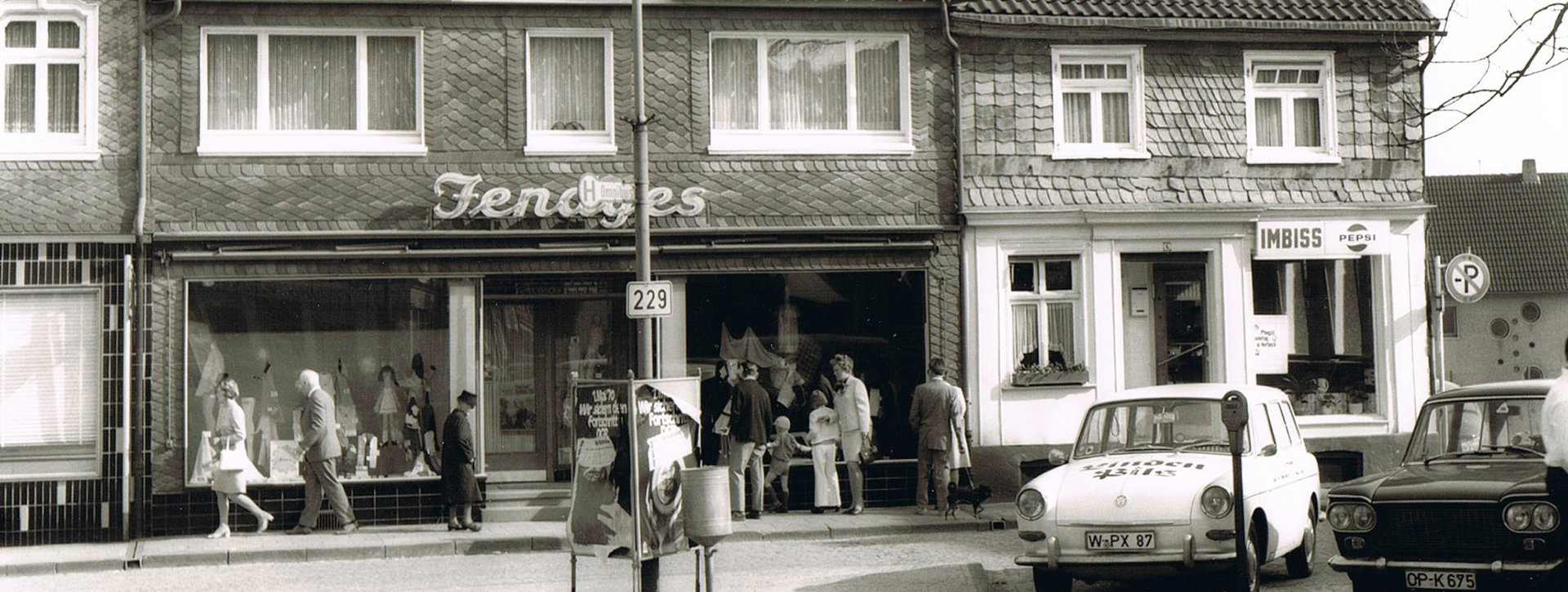 Schwarz-Weiß Foto aus den 1960er Jahren: Blick auf ein Geschäft, davor Fußgänger und parkende Autos