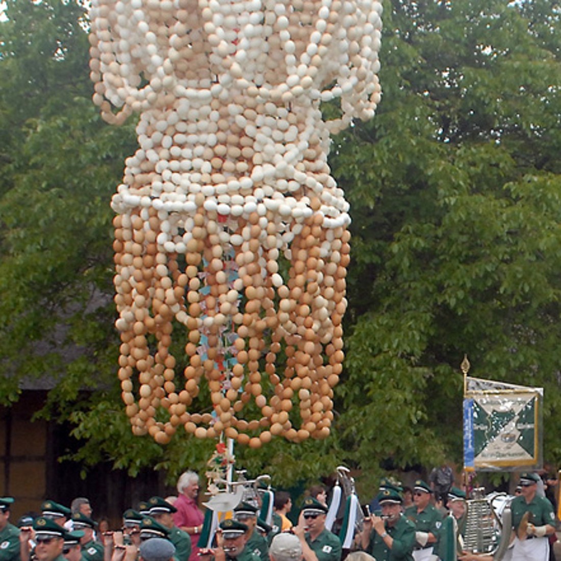 Ein Festumzug zieht an einem Fachwerkhaus vorbei, angeführt von einer Musikkapelle in grüner Uniform. Über dem Weg hängt eine große Krone aus Eiern