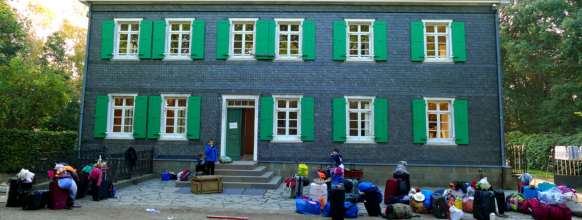 Graues Haus mit grünen Fensterläden, davor Kinder mit Rucksäcken