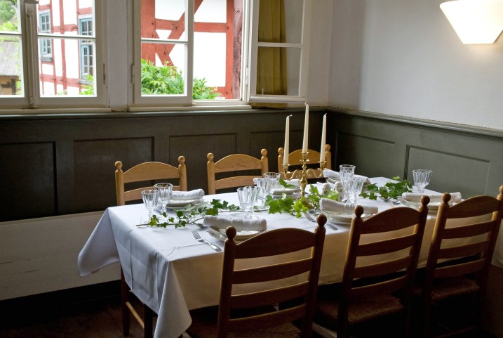 Tisch mit weißer Tischdecke, weißen Tellern, Kristallgläsern, grünen Blättern als Decke. Der Blick geht nach draußen auf einen Fachwerkwand