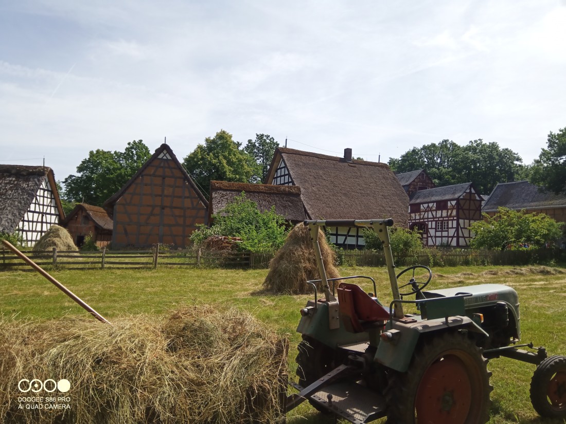 Woman sitting outdoors in front of a half-timbered house, spinning wool with a spinning wheel