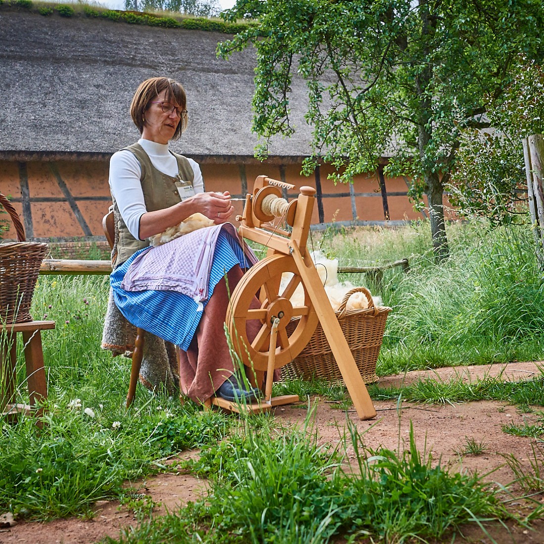 Frau sitzt im Freien vor einem Fachwerkhaus und spinnt Wolle mit einem Spinnrad