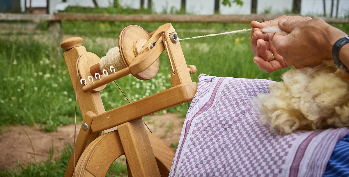 Woman sits outdoors in front of a half-timbered house and spins wool with a spinning wheel
