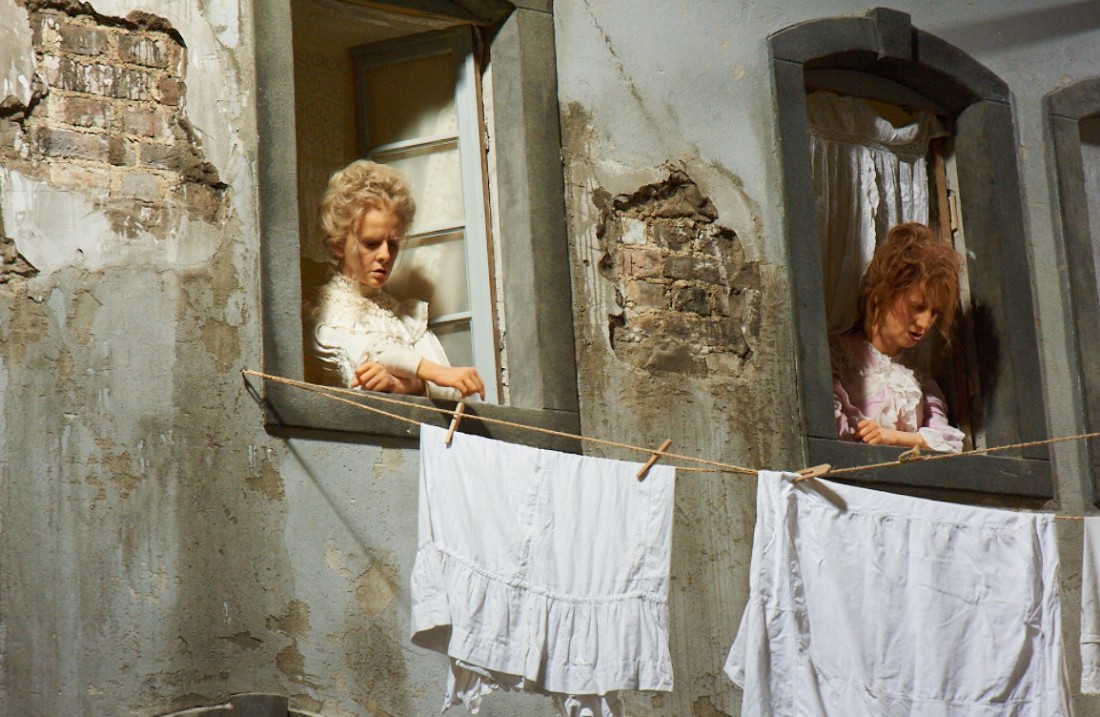 Scene from the exhibition, you see two women looking out of the windows of a dilapidated house, in front of which is a clothesline with white laundry stretched
