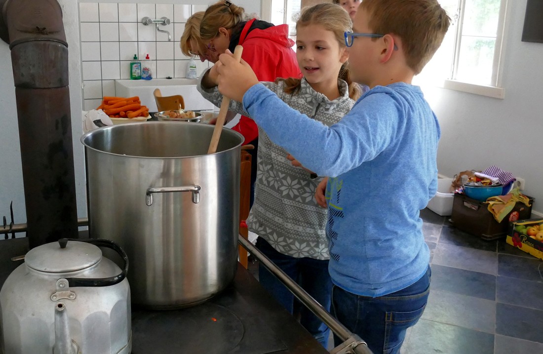 A boy stirs in a large pot with a wooden spoon, a girl helps him