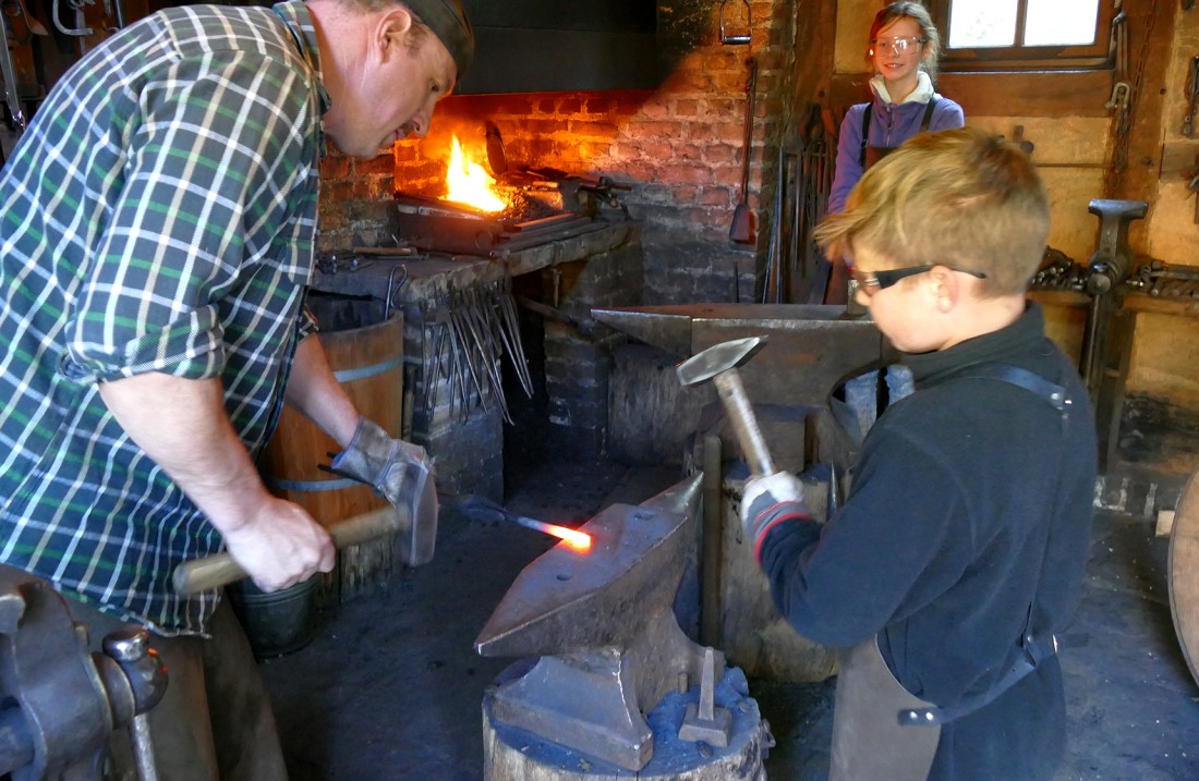 A blacksmith shows a boy how he processes glowing iron